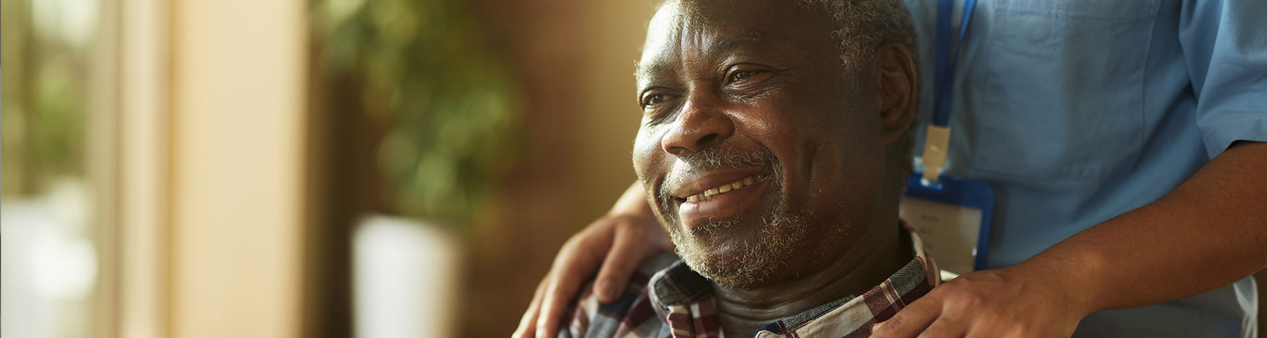 An elderly man with a warm smile sits near a window, with a caregiver in a blue uniform gently placing hands on his shoulders.