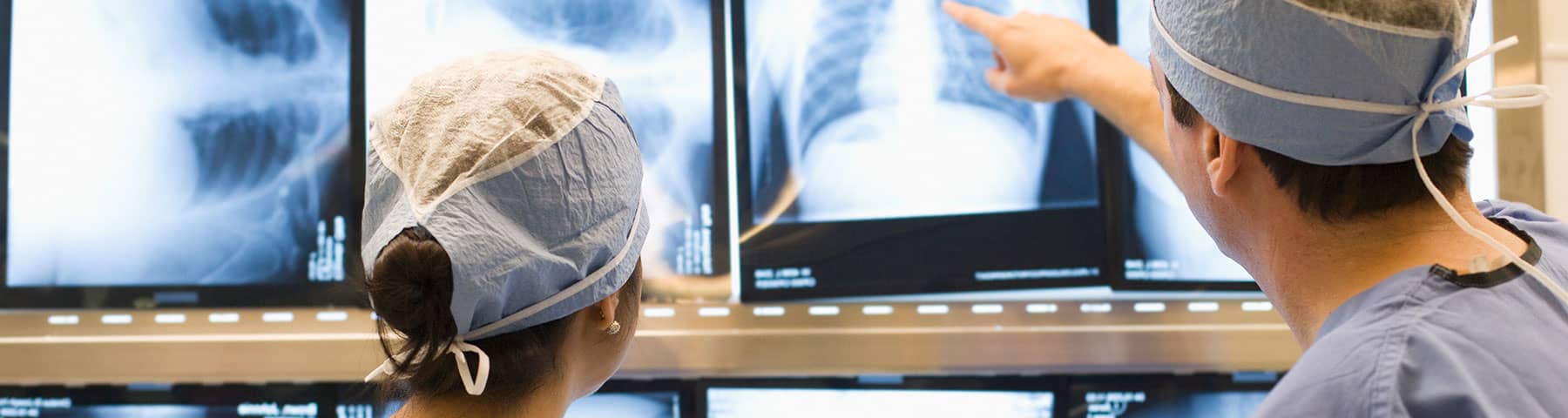 Two surgeons wearing blue scrubs and surgical caps analyze a series of X-ray images displayed on a screen, with one pointing towards a specific area.