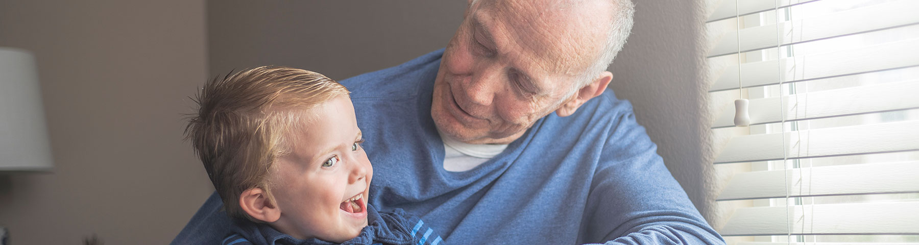 An elderly man in a blue sweater smiles warmly at a young child in his arms as they share a joyful moment near a bright window.