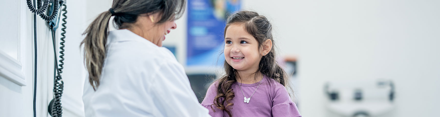 A young girl in a purple shirt smiles brightly while sitting in a doctor's office, engaging with a friendly female physician in a white coat.